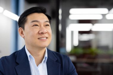 Hayalperest ve güler yüzlü adam, Asyalı iş adamı ofiste takım elbiseli ve düşünceli bir şekilde yan tarafa bakıyor, dinleniyor. Yakın plan fotoğraf..