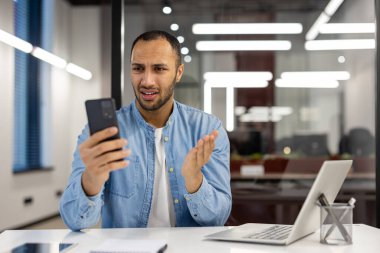 Genç bir Afrikalı Amerikalı ofiste çalışıyor. Masada endişeli bir şekilde oturuyor ve hayal kırıklığı ve şok içinde telefon ekranına bakıyor..