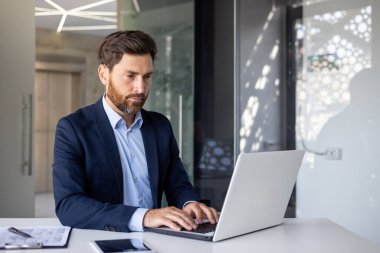 Konsantre olmuş genç ofis çalışanı takım elbiseli bir masada oturuyor ve dizüstü bilgisayarın klavyesine yazıyor..