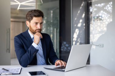 Ciddi ve düşünceli genç bir iş adamı masadaki ofiste oturuyor ve dizüstü bilgisayarına konsantre oluyor..