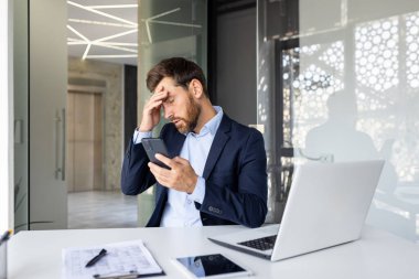 Şok olmuş genç işadamı, ofiste bir sandalyede oturuyor ve eliyle başını tutuyor, telefonu tutuyor, aldığı haberden rahatsız oluyor..