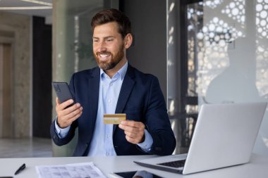 Başarılı finansal işlemler ve büyüme. Gülümseyen ve tatmin olmuş genç bir işadamı ofisteki masada oturuyor, elinde bir banka kartı tutuyor ve telefon ekranına bakıyor..