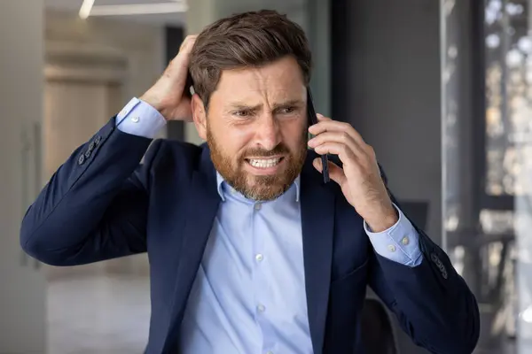 Close-up photo of a young businessman man in a suit standing in an office center, worriedly talking on the phone and holding his head with his hand in frustration, solving financial problems.