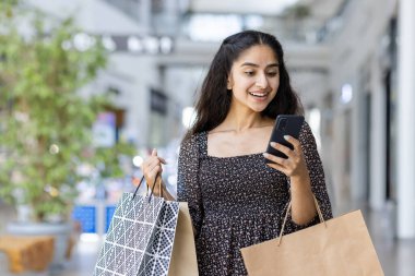 Mutlu genç bir Hintli kadın alışveriş merkezinde alışveriş yapıyor, içeride duruyor, elinde kağıt torbalarla cep telefonu kullanıyor..