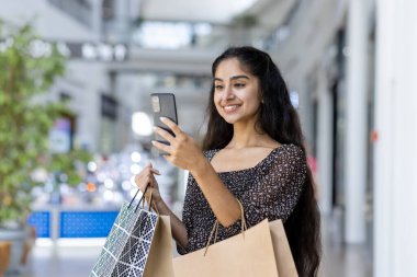 Hintli genç kız selfie çekiyor, video görüşmesinde konuşuyor, alışveriş merkezinin içinde blog yazıyor, alışveriş torbalarıyla duruyor ve telefon kamerasına bakıyor..