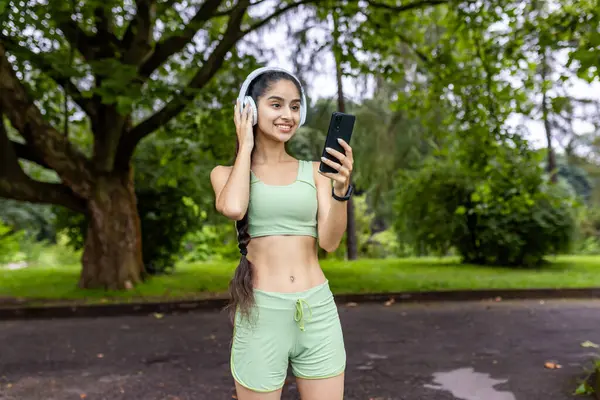 Young active Indian woman in green sportswear standing in park wearing headphones and looking at phone screen, jogging and exercising.