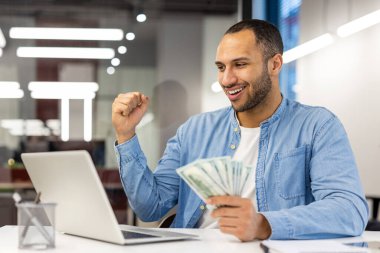 Dizüstü bilgisayarlı mutlu bir işadamı elinde para tutarken sevinçle finansal başarısını kutluyor..