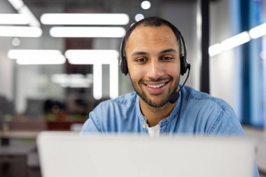 Neşeli bir erkek müşteri hizmetleri temsilcisi mikrofonlu bir kulaklık takıyor, çağdaş ofis ortamında bir bilgisayarda çalışıyor..