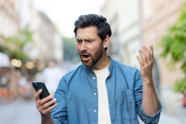 Sıradan giysiler içinde öfkeli bir genç adam bir şehir caddesinde duruyor ve endişeli bir şekilde telefon ekranına bakıyor, ellerini tutarsız bir şekilde sallıyor..