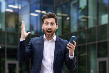Elinde telefon tutan genç bir işadamının yakın plan portresi. Bir ofis binasının önünde duruyor. Şok ve şaşkınlık içinde kameraya bakıyor..
