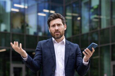 Hayal kırıklığına uğramış genç bir iş adamının yakın plan portresi bir ofis binasının yanında duruyor, elinde bir telefon tutuyor ve açıklanamaz bir şekilde kameraya kollarını sallıyor..