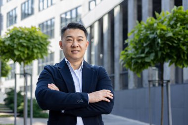 Gülümseyen Asyalı işadamı takım elbiseli, kollarını kavuşturmuş, kendine güvenen ve profesyonelce kameraya bakıyor..