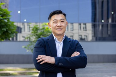 Takım elbiseli profesyonel bir işadamı modern bir ofis binasının önünde kollarını kavuşturarak başarıyı somutlaştırıyor..