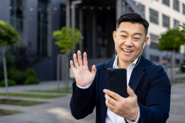 Bir video görüşmesi yapan mutlu profesyonel bir adam, kulaklıklı meslektaşlarına el sallıyor ve dışarıda gülümsüyor..