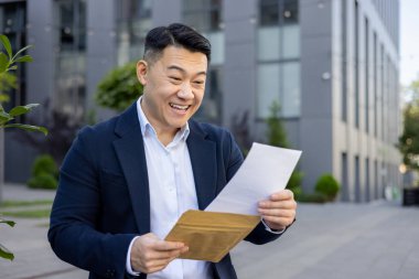 Mutlu Asyalı işadamı ofis binasının dışında heyecan dolu bir mektup okuyarak başarı ve neşeyi ifade ediyor..