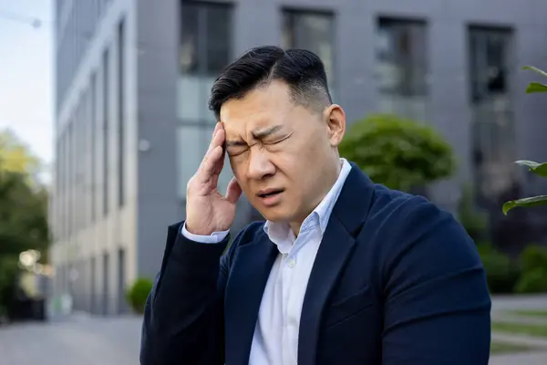 Worried Asian businessman in a blue suit feeling a headache, standing outside an office building with a pained expression.