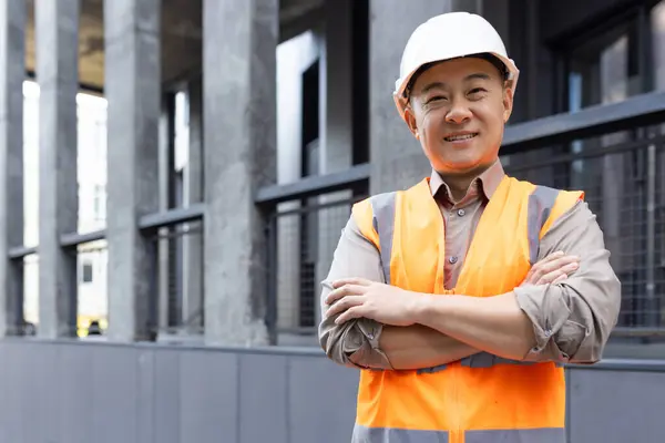 Portrait of a happy male construction worker with crossed arms wearing a safety helmet and reflective vest at a construction site.