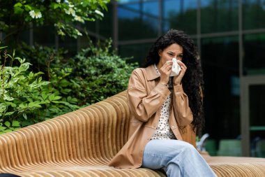 A woman sits on an outdoor bench, blowing her nose with a tissue, possibly suffering from cold or allergies, in a natural, urban setting. clipart