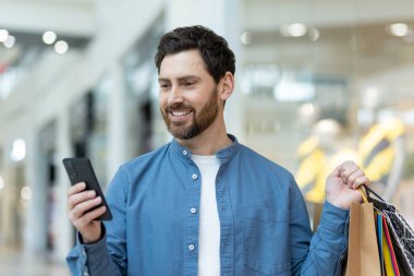 Renkli çantaları olan genç bir erkek alışveriş merkezinin içinde telefonuna bakıyor..