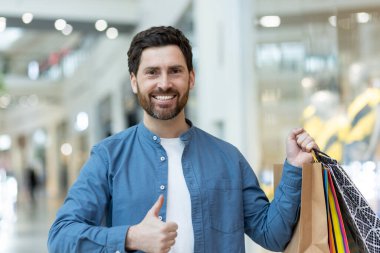 Parlak bir alışveriş merkezinde poşet tutarken, rahat gömlekli bir adam baş parmak işareti yapıyor..