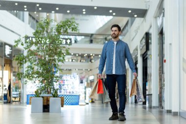 Gülümseyen genç bir adam alışverişinden memnun bir şekilde bir alışveriş merkezinde birden fazla renkli çantayla yürüyor..
