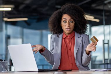 Afro-Amerikalı bir kadının elinde dizüstü bilgisayarla otururken kredi kartıyla el sallaması şok edici. Kafası karışmış ofis çalışanı, kabinedeki zayıf bağlantı nedeniyle çevrimiçi işlemle mücadele ediyor.