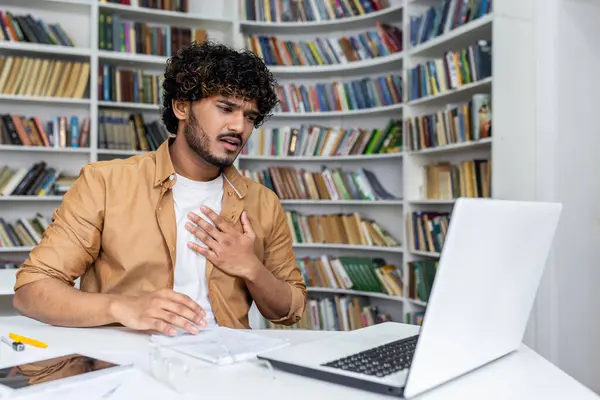 Millennial kıvırcık tişörtlü adam masaüstünde portatif bilgisayarla oturuyor ve elini göğsüne koyuyor. Stresli muhasebeci kalp krizi belirtileri gösteriyor ve iş yerinde nefes almakta güçlük çekiyor..