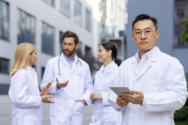 Serious chinese doctor looking at camera and holding digital tablet while standing on blurred background with colleagues. Group of medical workers in while lab coats discussing new treatment strategy.