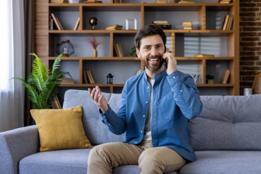 Neşeli sakallı adam cep telefonuyla sohbet ediyor, modern oturma odasındaki koltukta rahatça oturuyor..