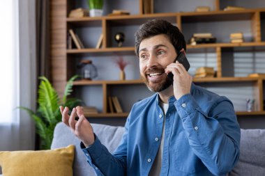 Mavi gömlekli sıradan bir adam modern bir oturma odasında cep telefonuyla konuşurken el kol hareketi yapıyor..