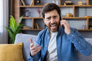 Modern bir oturma odasında stresli bir telefon görüşmesi yapan yetişkin bir adam üzgün görünüyor..