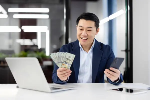 Professional businessman in a suit displaying cash and using a smartphone at a modern office work station.