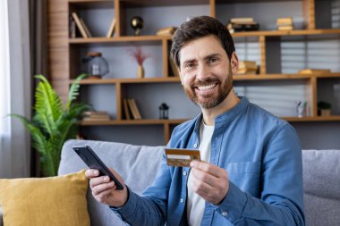 Kanepede rahat rahat oturan, akıllı telefon kullanan ve rahat bir oturma odasında online işlemler için kredi kartı tutan sıradan bir erkek..