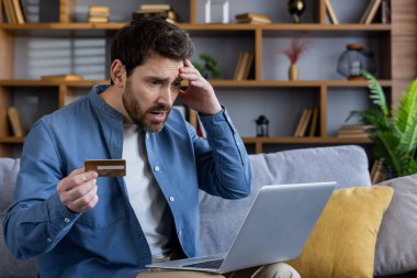 Kredi kartı tutan endişeli bir erkek bilgisayarına şokla bakıyor, muhtemelen online dolandırıcılık ya da bankacılık sorunlarıyla karşı karşıya..