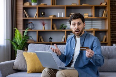 Şok olmuş genç bir erkek elinde kredi kartıyla koltukta oturuyor ve şaşkın bir ifadeyle dizüstü bilgisayar kullanıyor..