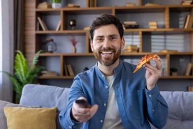 Neşeli orta yaşlı bir adam elinde pizza dilimi ve uzaktan kumandayla oturma odasındaki kanepede mutlu bir ifadeyle dinleniyor..