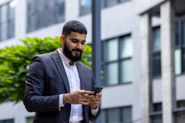 Takım elbiseli iyi giyimli bir iş adamı akıllı telefonuyla çağdaş bir ofis ortamının dışında meşgul olur. Şirket ortamında iletişim ve teknolojiye odaklan.