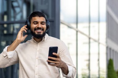 Genç bir Hintli adam sokakta gömlek ve kulaklıkla duruyor, elinde bir telefon tutuyor, ekrana bakıyor, konuşuyor, müzik dinliyor, dans ediyor..