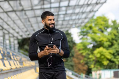 Spor kıyafetleri ve kulaklık takan, gülümseyen, sakallı bir adam açık hava stadyumunda dururken akıllı bir telefon tutuyor..