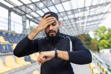 Yorgun bir erkek sporcu, yorgunluk ve kişisel fitness takibini gösteren akıllı hareketlerine bakarken alnından teri siler..