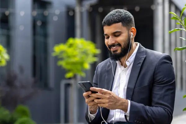 A modern businessman in a suit and earphones is engaged with his smartphone, smiling as he reads the screen, amidst an urban backdrop with verdant greenery.