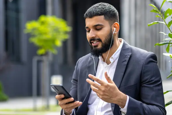 A well-dressed man engages with his smartphone while using earphones outside a modern building, possibly during a work break or in transit.