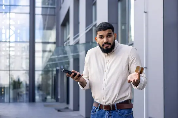Portrait of a young Arab man standing on the street near an office building, holding a credit card and a mobile phone, looking worriedly at the camera and spreading his hands.