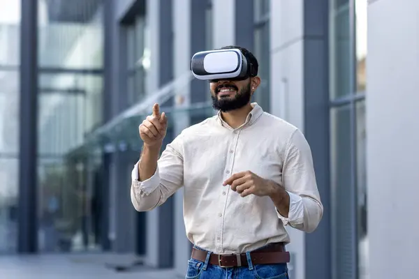 A young man with a beard is standing outside a modern building, wearing a virtual reality headset and gesturing with his hands as if interacting with the virtual environment. The image evokes a sense