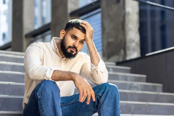 Upset and disappointed young Indian man sitting on the steps of an office building and holding his hand above his head, looking thoughtfully to the side.