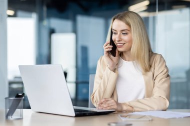 Bir iş kadını, modern ofis ortamında dizüstü bilgisayar kullanırken cep telefonuyla sohbet eder..