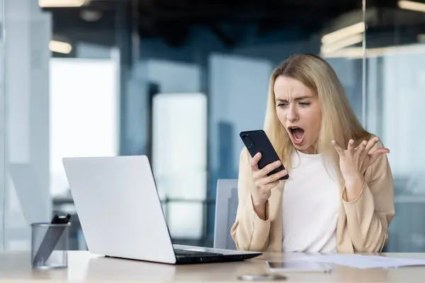 Angry professional woman showing frustration while looking at her smartphone at a modern office desk setup.
