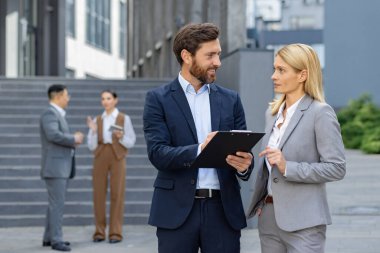 Takım elbiseli iki şirket çalışanı modern bir ofis binasının önünde bir panoyla tartışıyorlar..