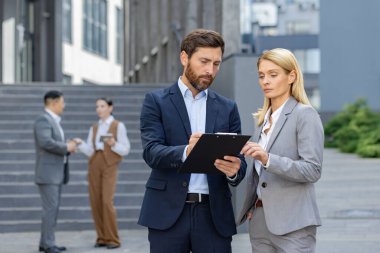 İki profesyonel iş adamı, arka planda meslektaşlarıyla birlikte ofis binasının dışında bir pano tutuyorlar..