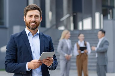 Arka planda bulanık meslektaşları olan bir tablet kullanan gülümseyen iş adamı, modern bir şirket ortamında liderlik ve teknoloji entegrasyonunu tasvir ediyor..